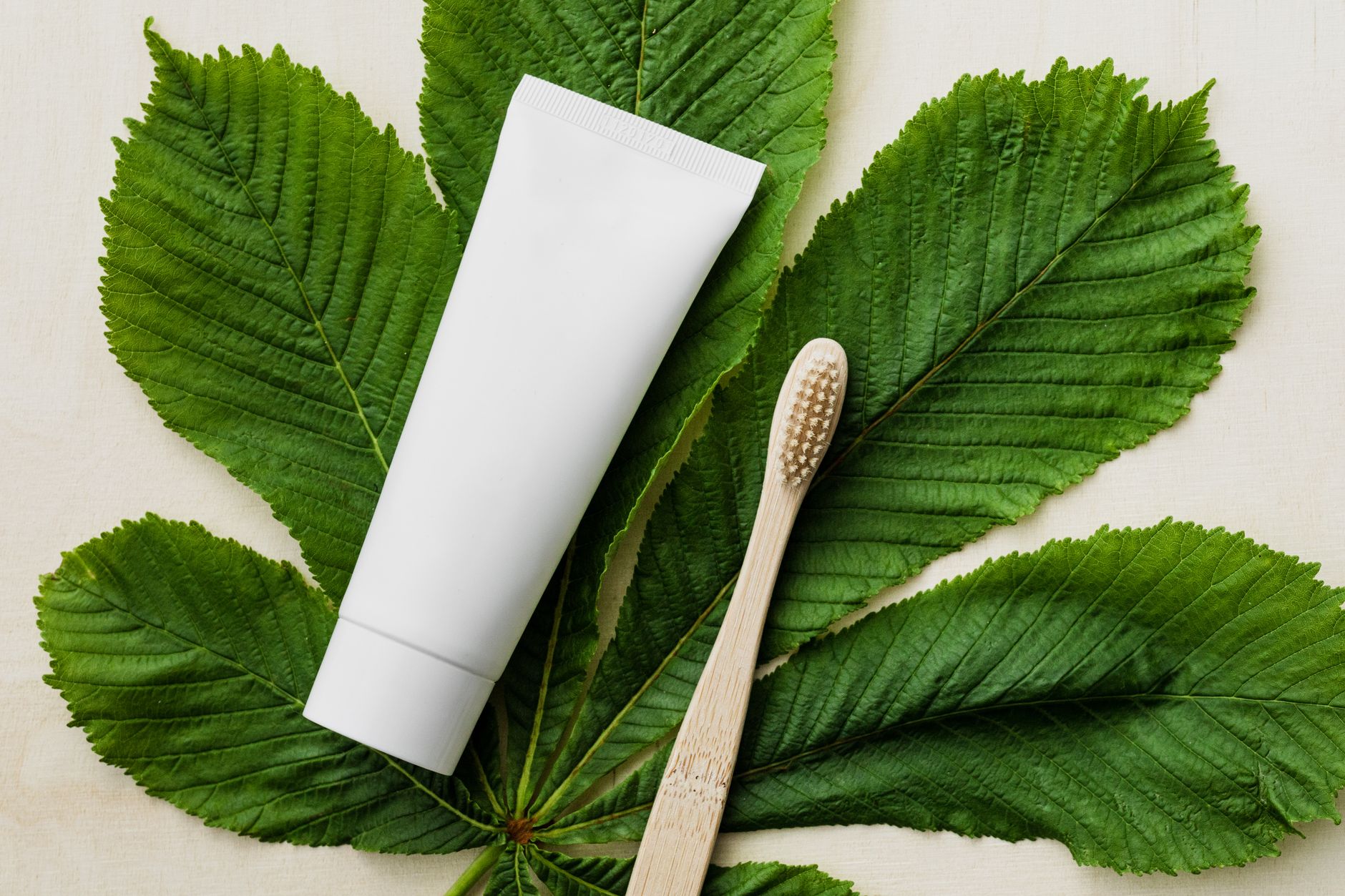 Tubo de pasta de dente branca com escova de dente de bambu em folhas verdes para cuidados dentais ecológicos.