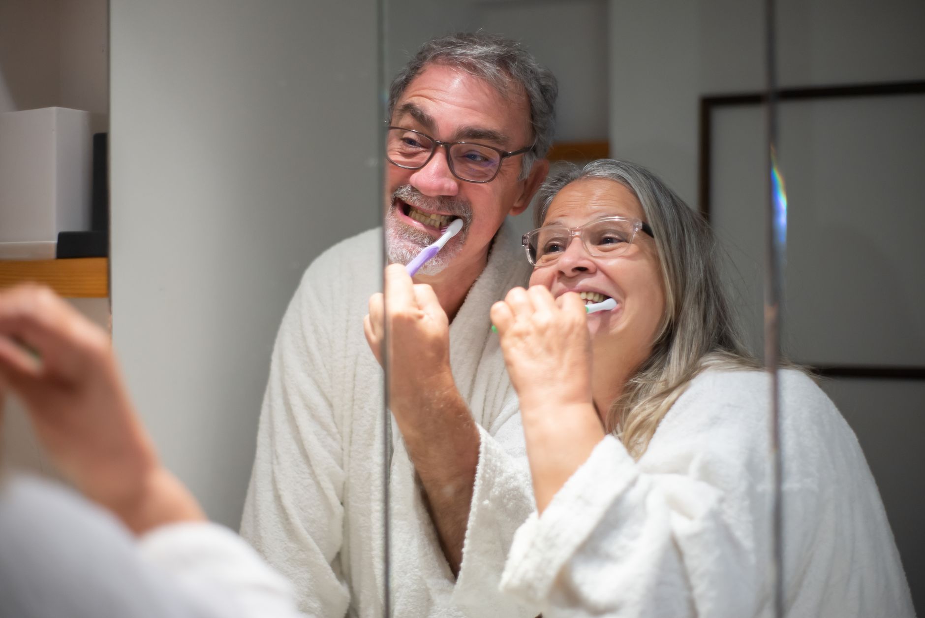 Casal feliz de idosos escovando os dentes juntos no espelho do banheiro, enfatizando a higiene dental.